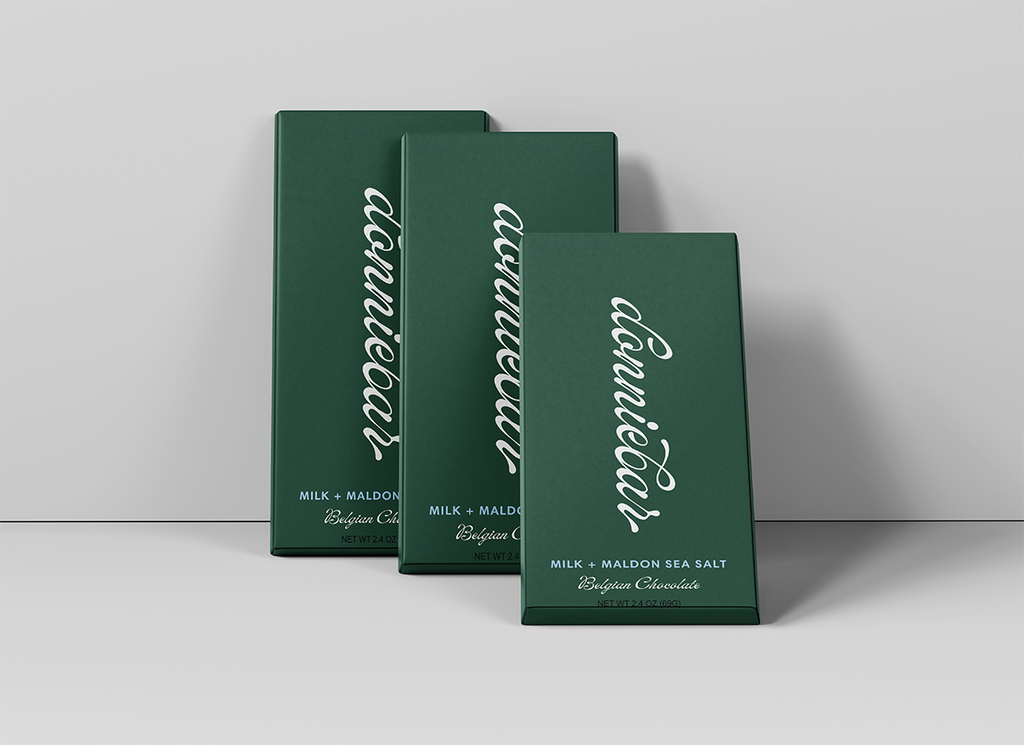 Three dark green rectangular chocolate boxes arranged in a staggered studio setup against a light gray background. The front box is slightly open, and all boxes feature large white cursive text reading 'conneault' with smaller white text below indicating milk and flavor details.