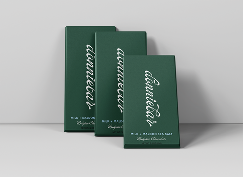 Three dark green rectangular chocolate boxes arranged in a staggered studio setup against a light gray background. The front box is slightly open, and all boxes feature large white cursive text reading 'conneault' with smaller white text below indicating milk and flavor details.