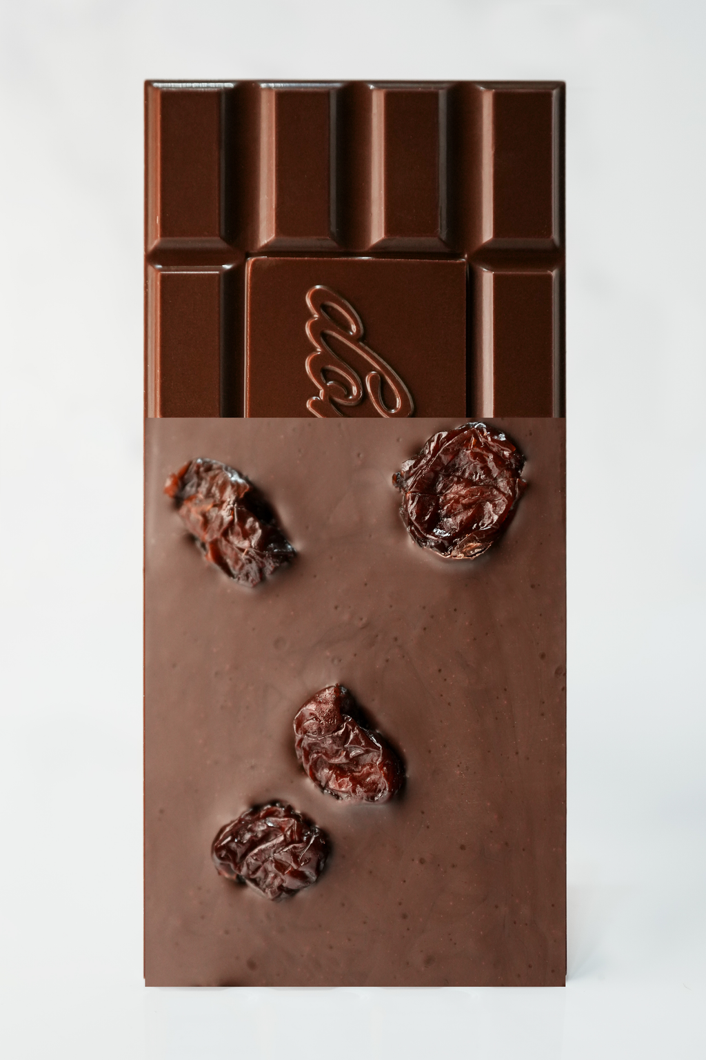 A chocolate bar with a matte milk-chocolate finish, featuring a decorative embossed logo in the upper center and three dried Morello cherries embedded vertically in the lower half, set against a clean white background.