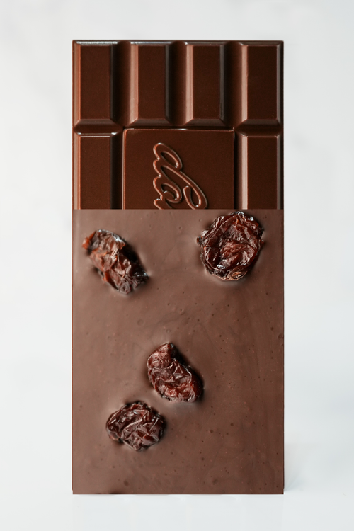 A chocolate bar with a matte milk-chocolate finish, featuring a decorative embossed logo in the upper center and three dried Morello cherries embedded vertically in the lower half, set against a clean white background.