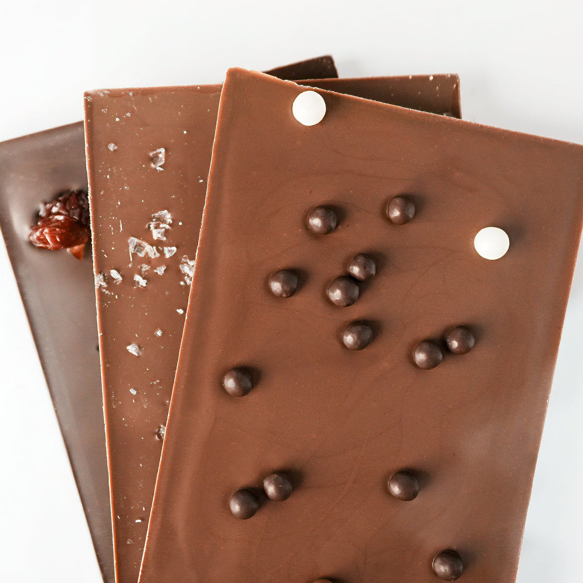 Three overlapping chocolate bars in different milk and dark chocolate varieties, displayed on a clean white background. The front bar is milk chocolate with chocolate chips, the middle is dark chocolate with sea salt flakes, and the back is a caramel chocolate piece.