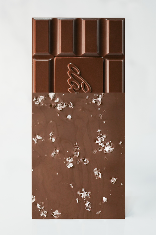 A vertical chocolate bar with a light brown upper section featuring a segmented pattern and a stylized embossed logo, and a lower section of milk-chocolate brown with scattered white sea salt flakes, set against a plain white background.